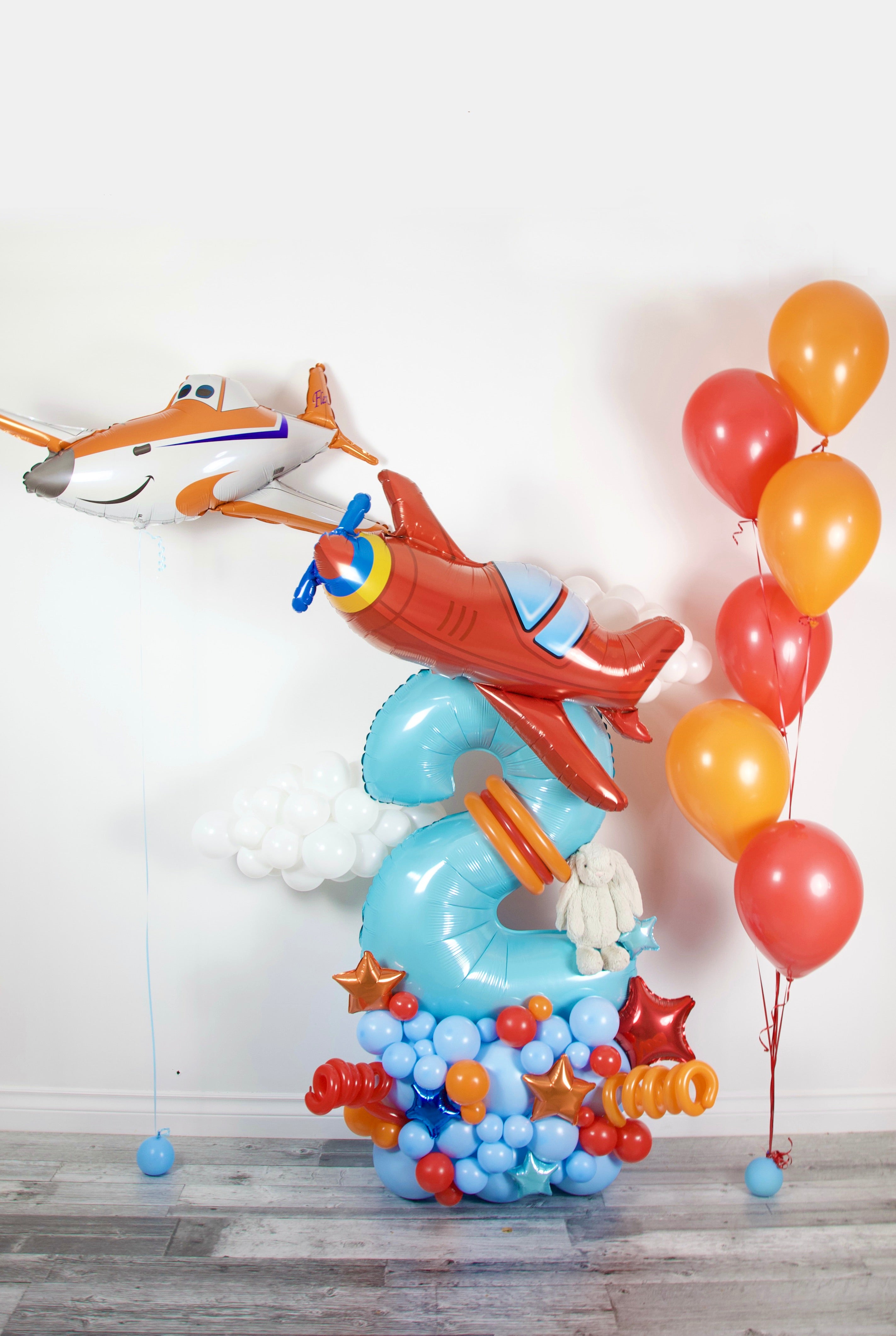 Balloon arrangement shaped like a number two with airplane design, accompanied by red and orange balloons on a light wooden floor.
