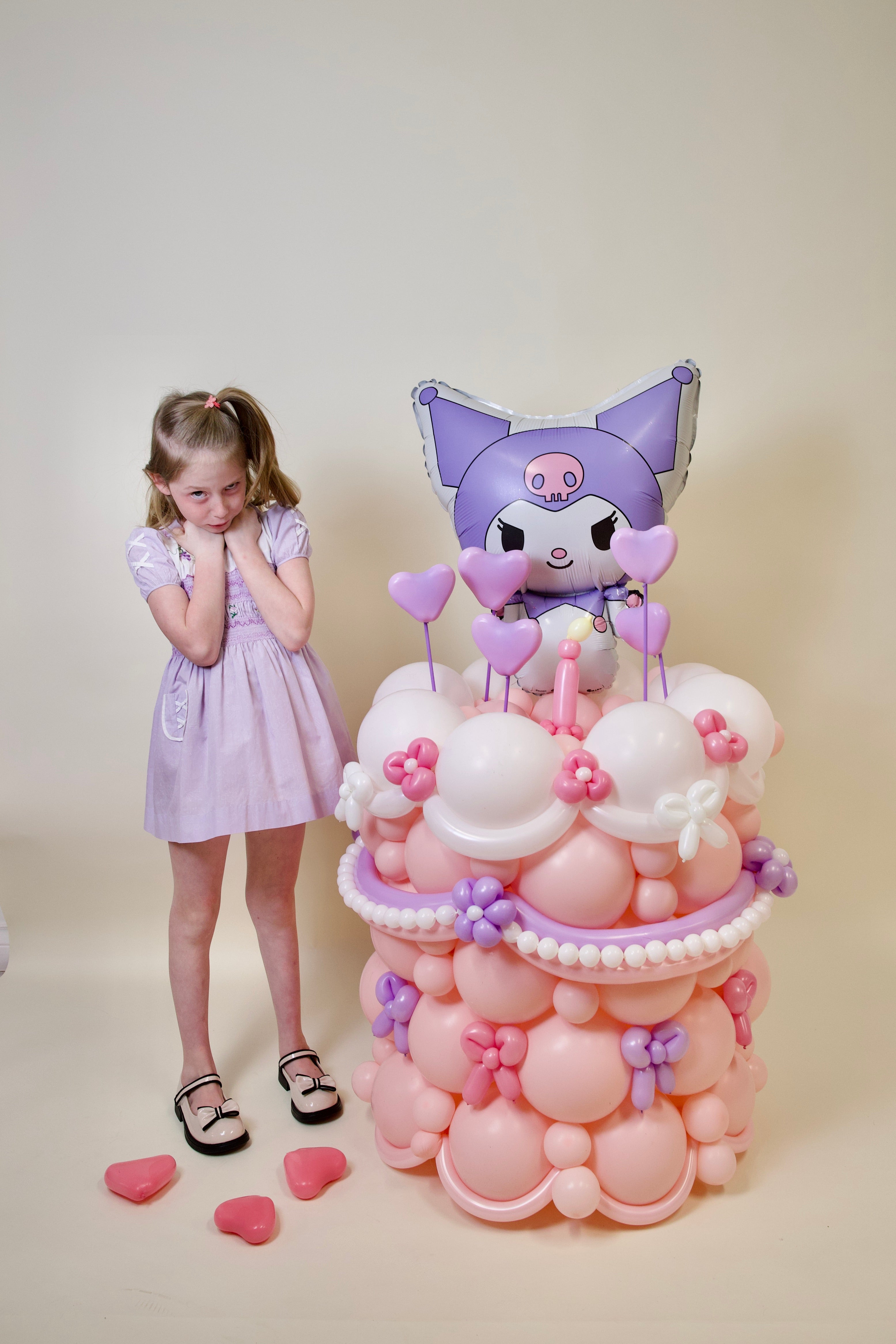 Child in a lavender dress standing next to a balloon sculpture with a character on a plain background