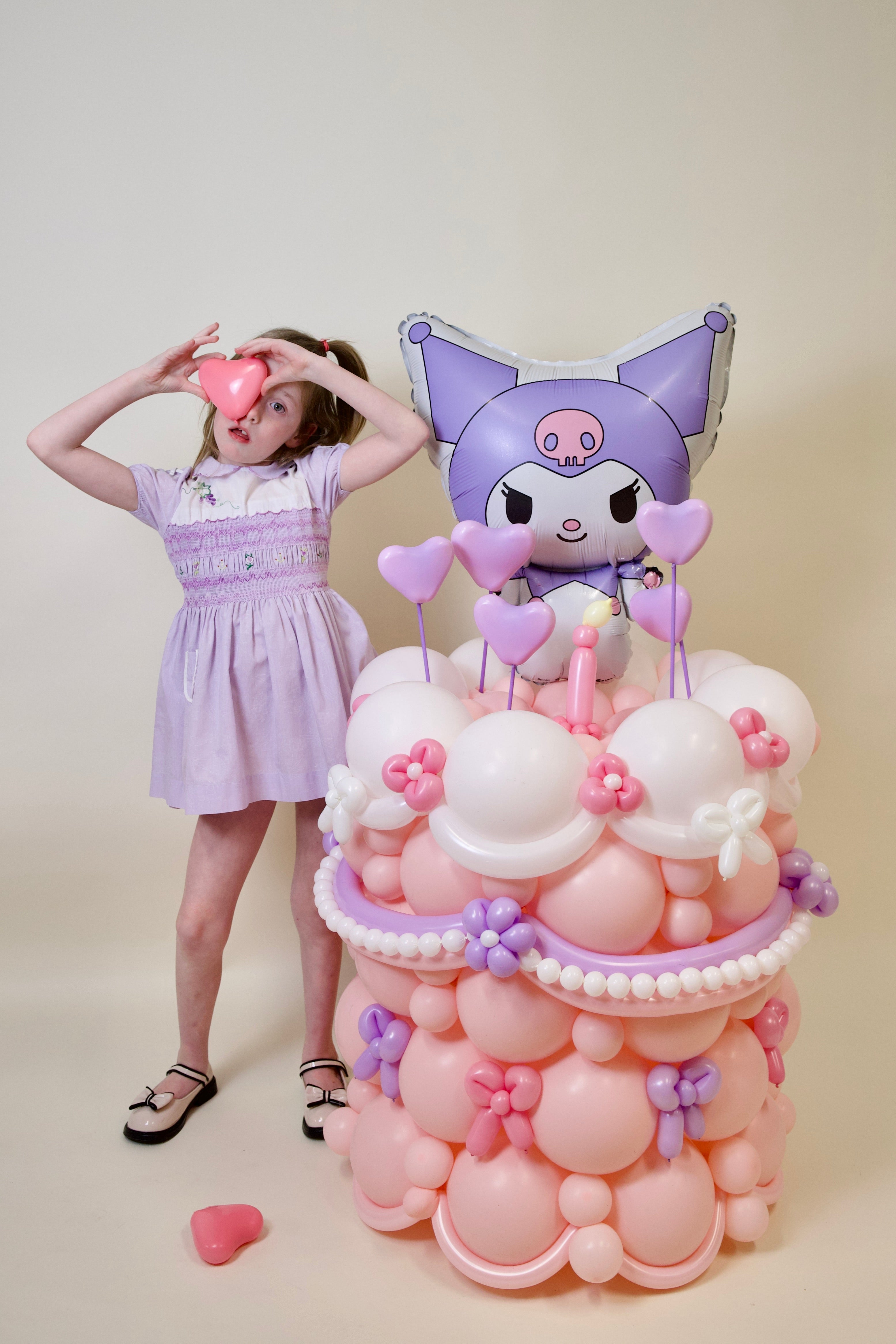 Girl in a lavender dress standing next to a balloon sculpture with a character on a beige background