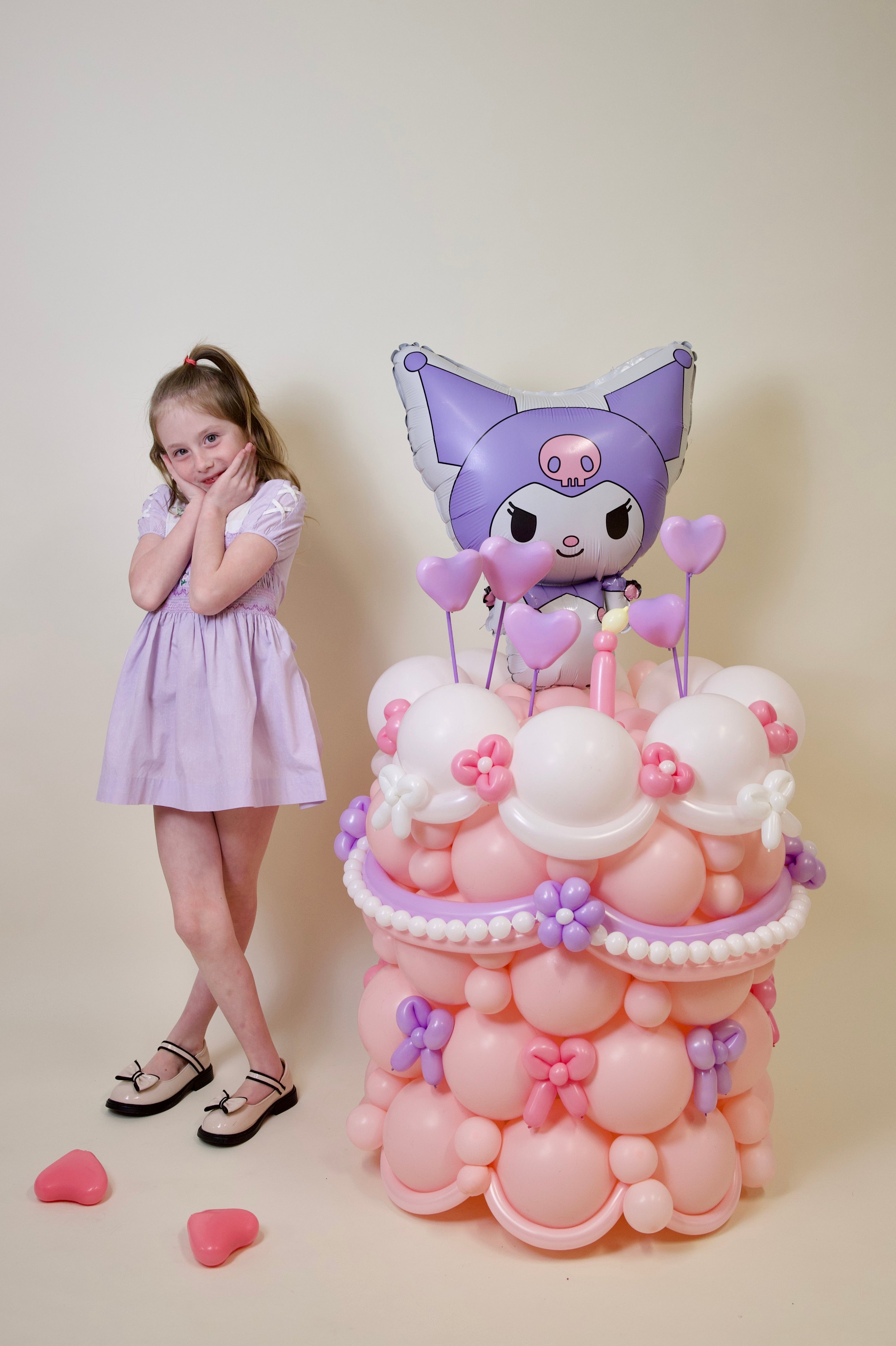 Girl in a light purple dress standing next to a balloon sculpture with a character on a beige background