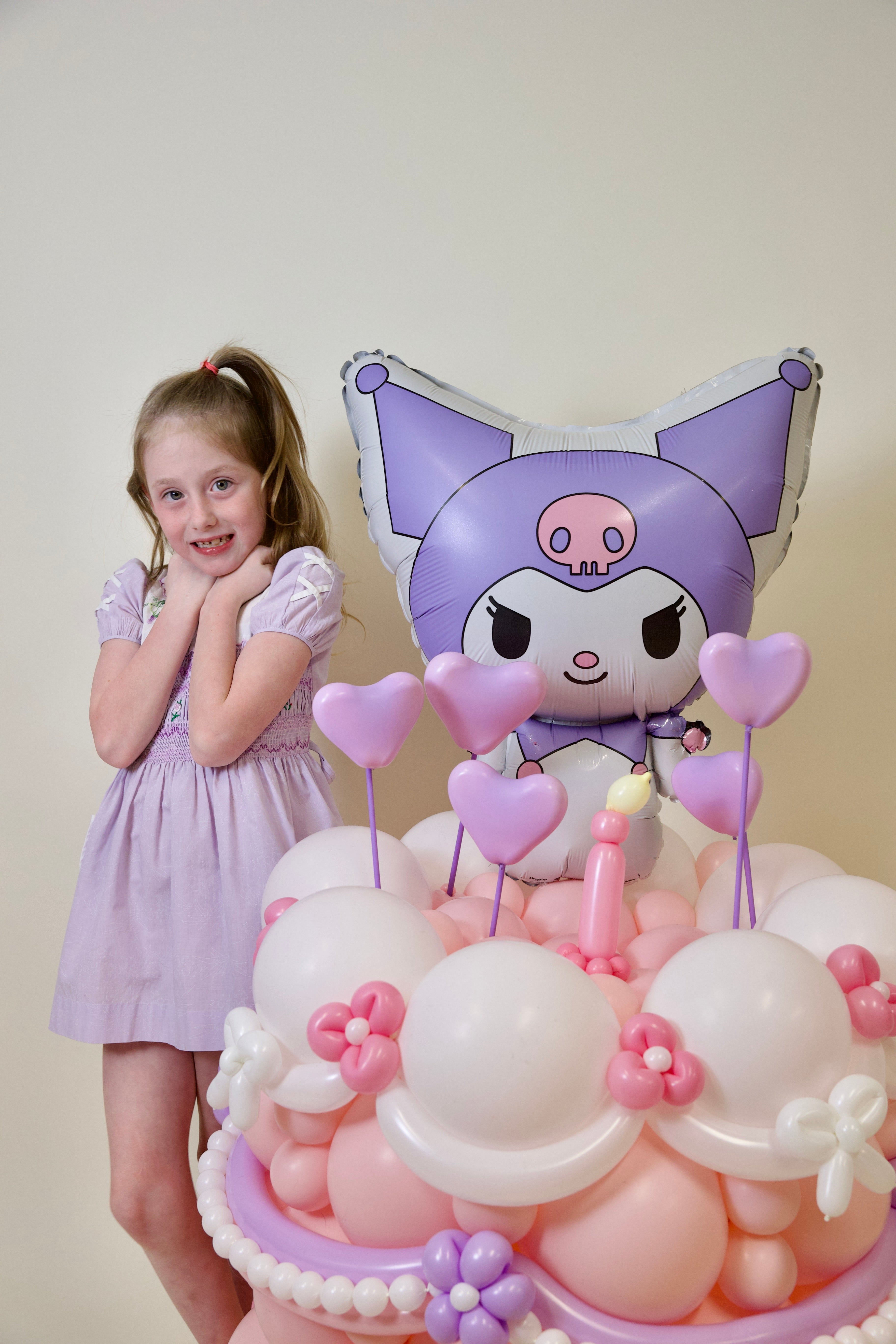 Child in a lavender dress standing next to a decorative cake with a cartoon character balloon.