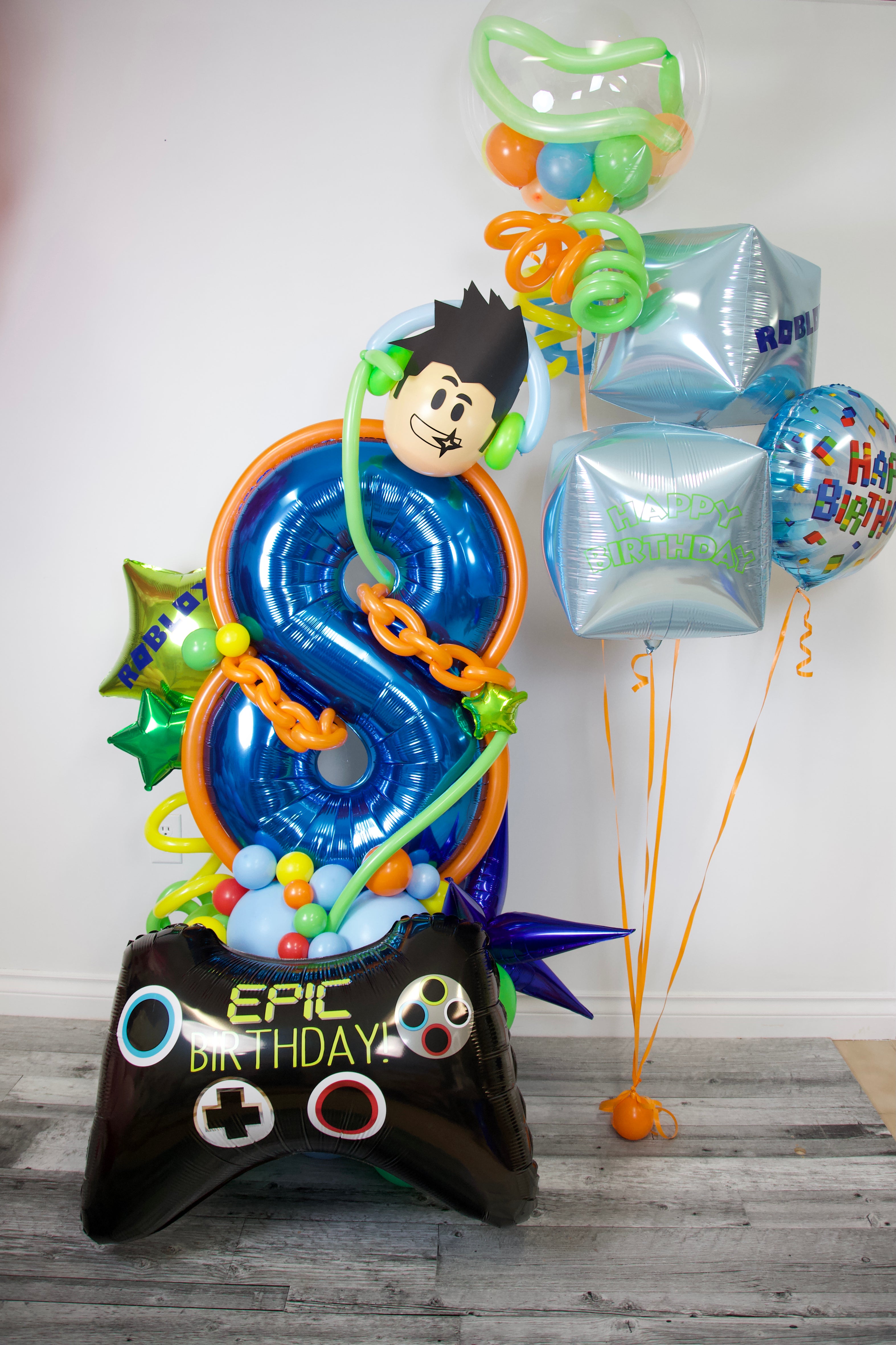 Colorful balloon arrangement with a large number 8 and 'Epic Birthday' controller, featuring a character face, on a wooden floor.