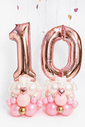 Rose gold number balloon columns with blush balloon bases, custom age digits, mini heart accents and floral details